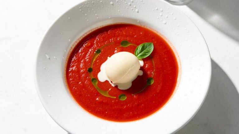 Chilled tomato soup in a white bowl with a scoop of parmesan ice cream melting in the center