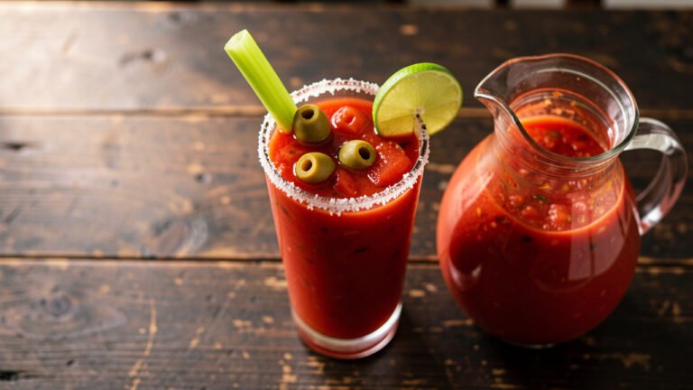 A tall glass of bloody mary made from homemade mix, garnished with celery, olives, and lime, with a pitcher of mix beside it